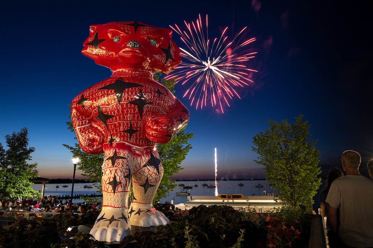 A red, star‑patterned Bucky Badger statue stands by a lakeshore as fireworks burst in the night sky.