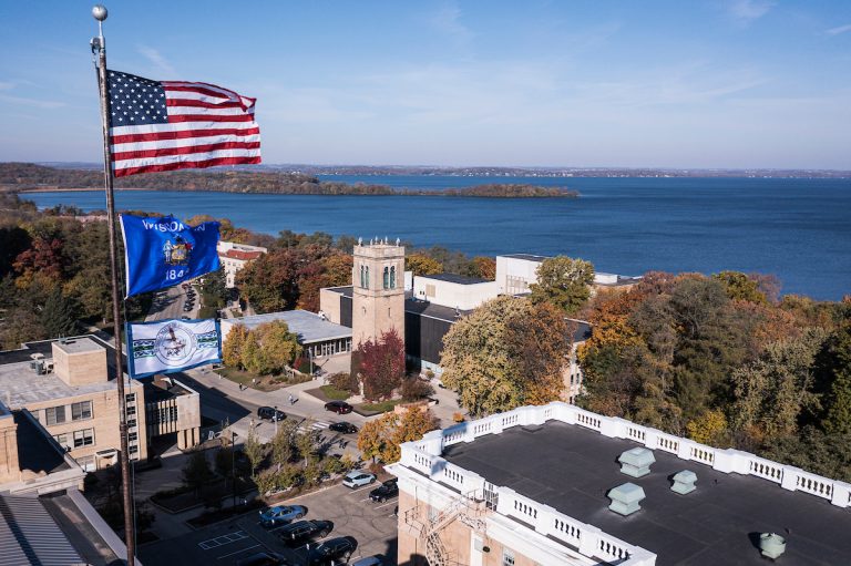 Aerial image of the US flag, Wisconsin State Flag and Ho-Chunk Nation flag flying over Bascom Hall with campus, Picnic Point and Lake Mendota pictured in the background.