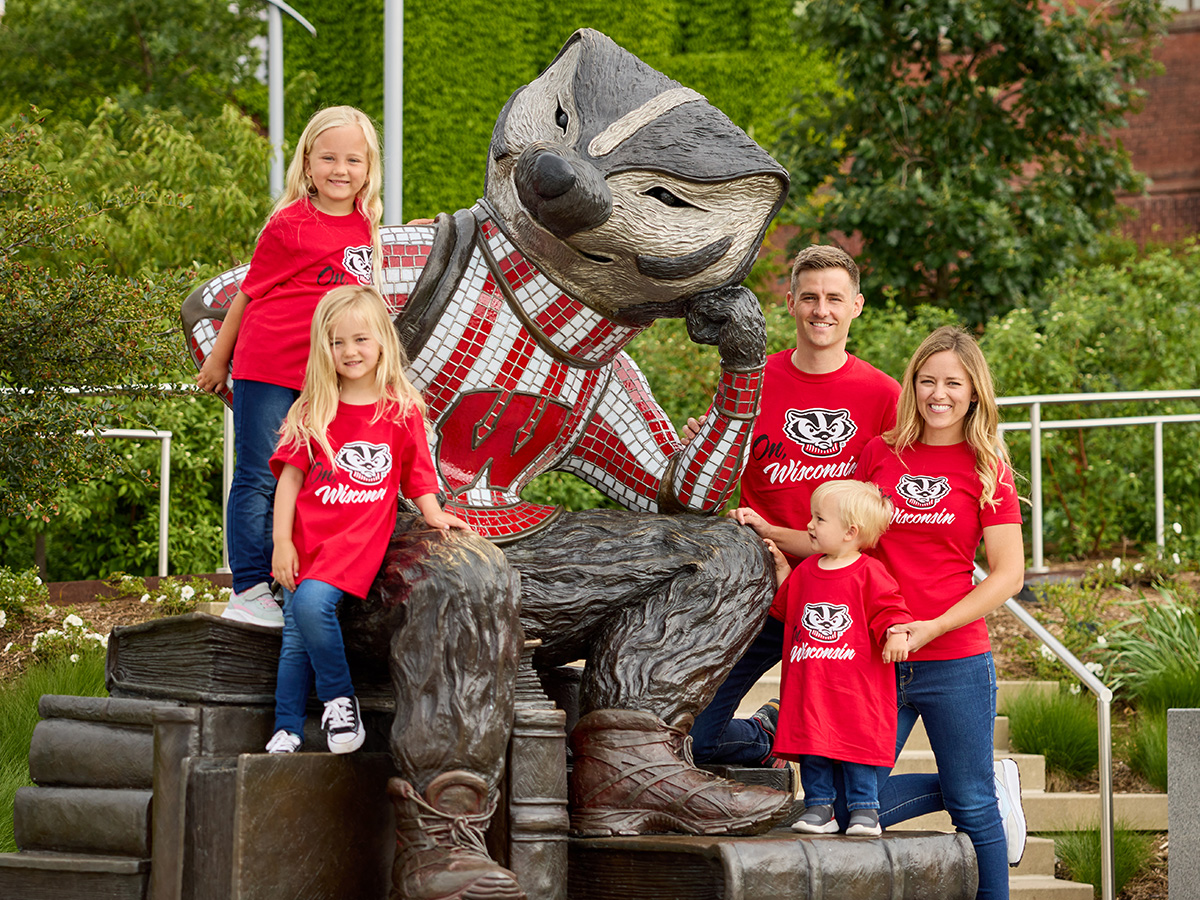 Two parents pose with their three children at Alumni Park, all wearing The Red Shirt, a red t-shirt with Bucky Badger's face on it and the words 'On Wisconsin'.