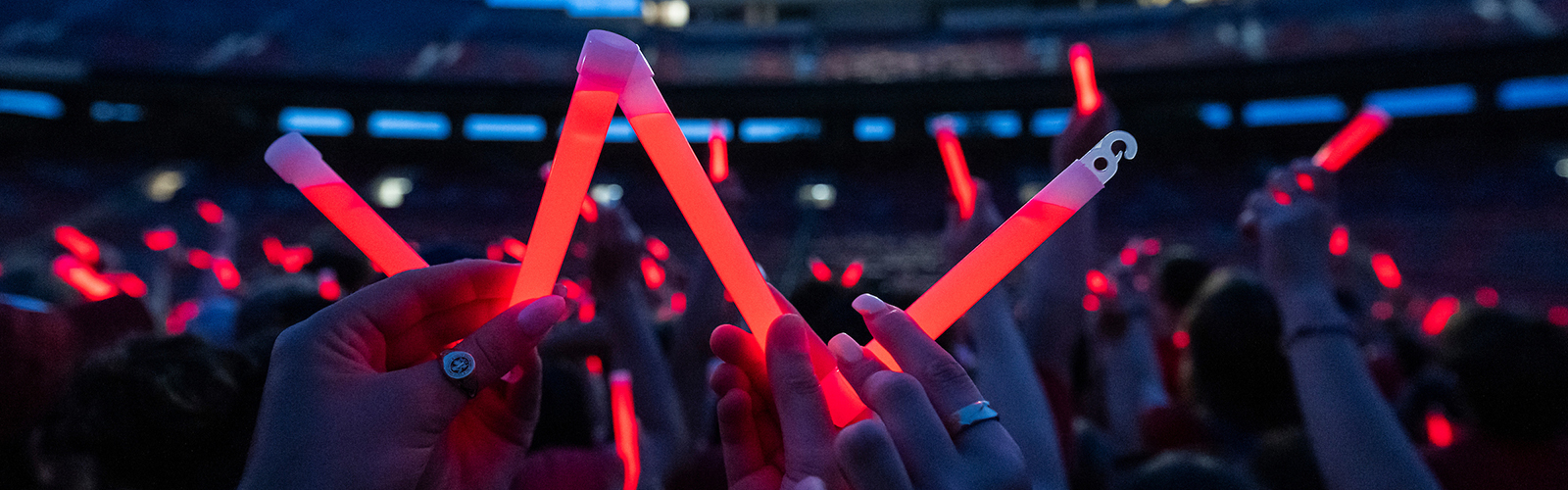 Crowd in dimly lit stadium holding up red glow sticks, with close-up of someone forming a "W" using the glow sticks.