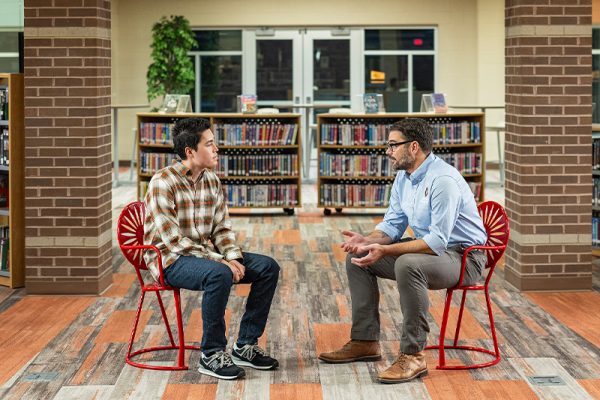 Two people sitting on red Wisconsin Union Sunburst Terrace chairs, talking face-to-face and surrounded by books in a library.