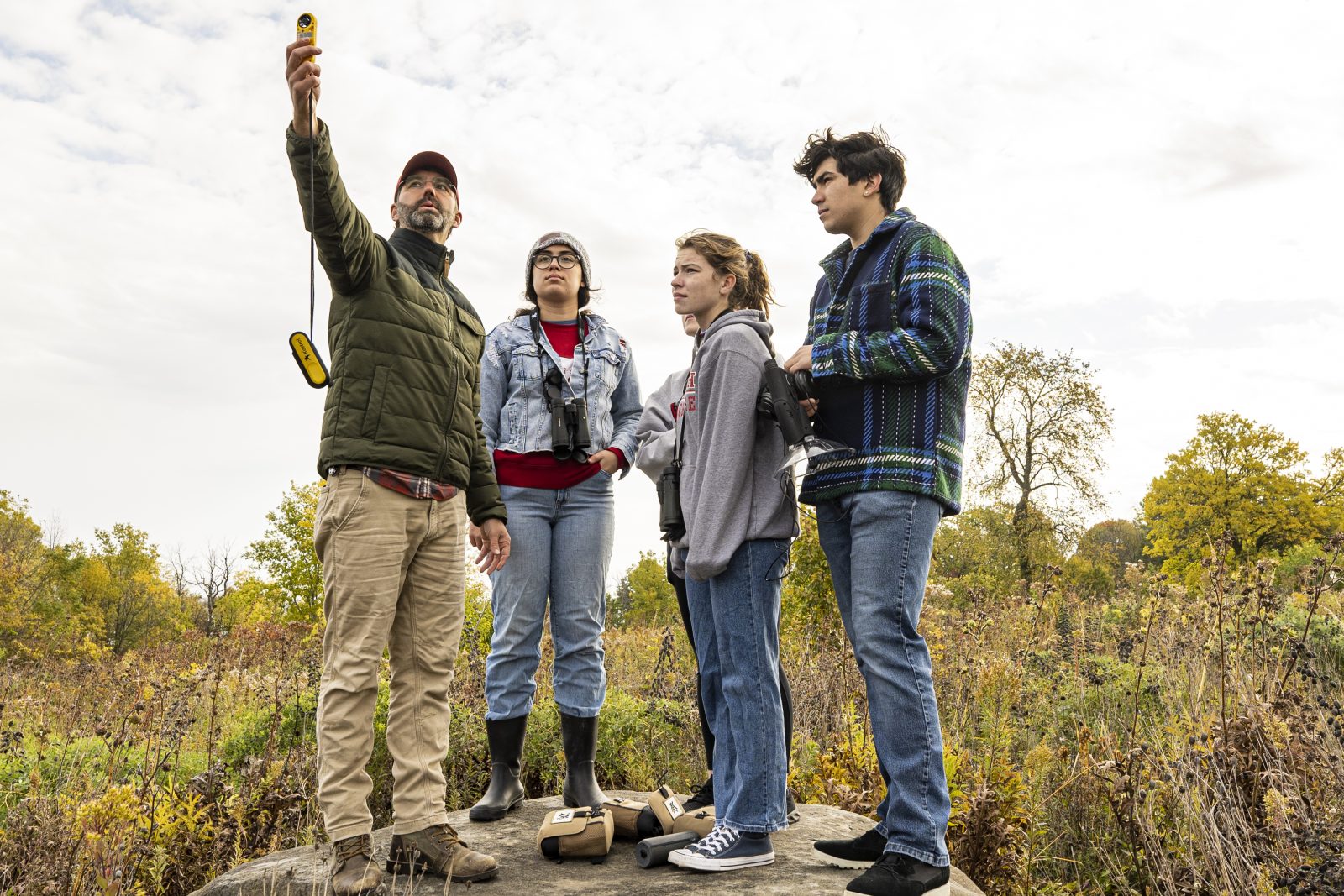 Four people standing on rock in prairie holding binoculars and yellow handheld device, collecting research data on bird populations.