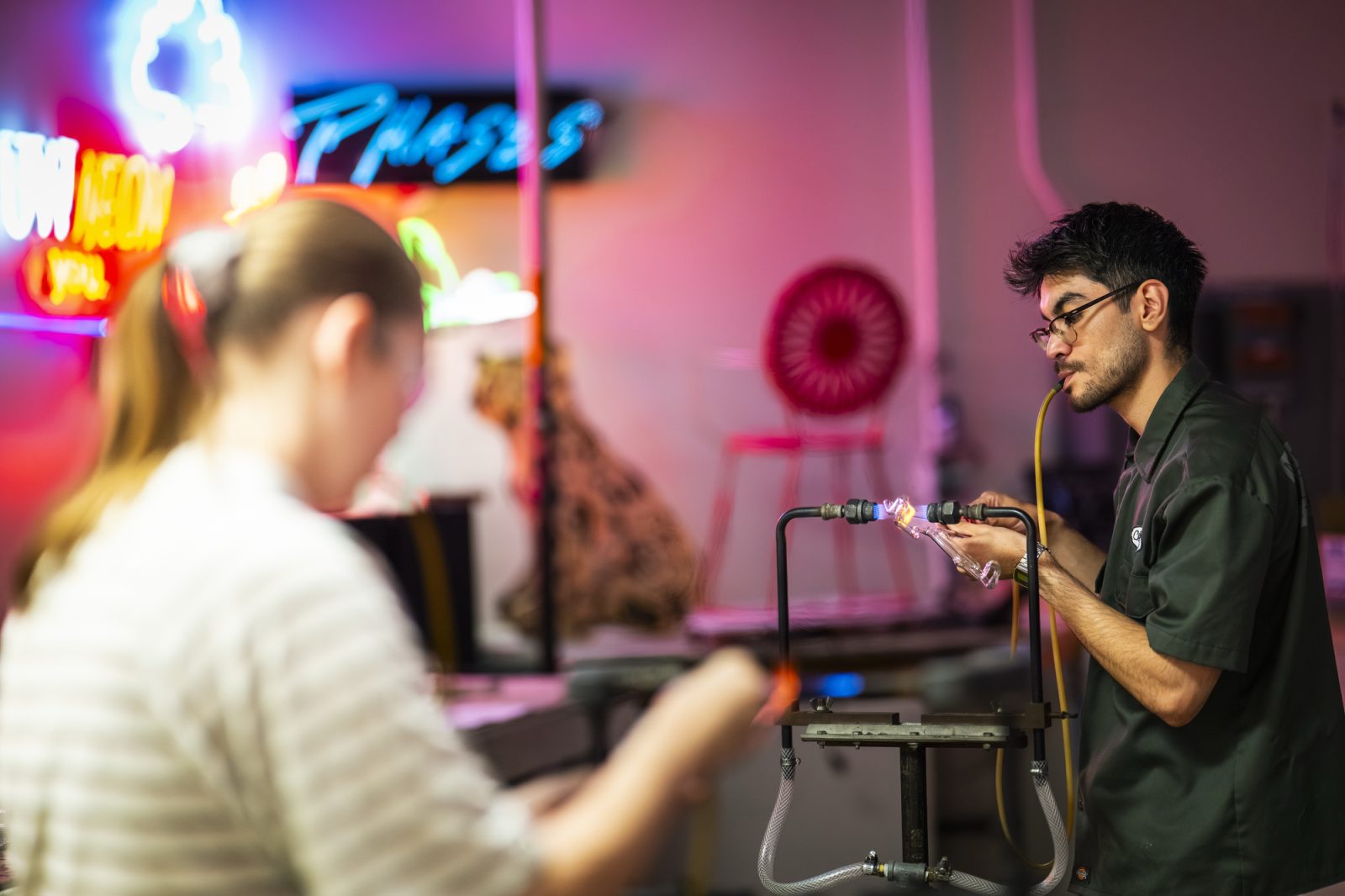Person glassblowing in vibrant studio with neon signs and eclectic decor.