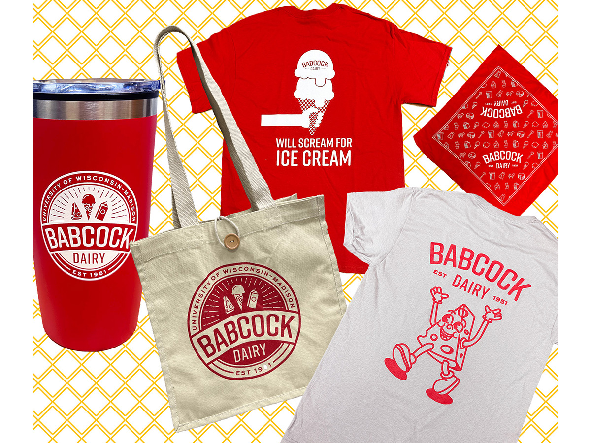 A collection of Babcock Dairy Store merchandise including a red travel tumbler, a beige tote bag, a red T-shirt with an ice cream graphic and the text ‘Will Scream for Ice Cream,’ a red bandana, and a light gray T-shirt with a cheese cartoon character, all featuring Babcock Dairy branding.