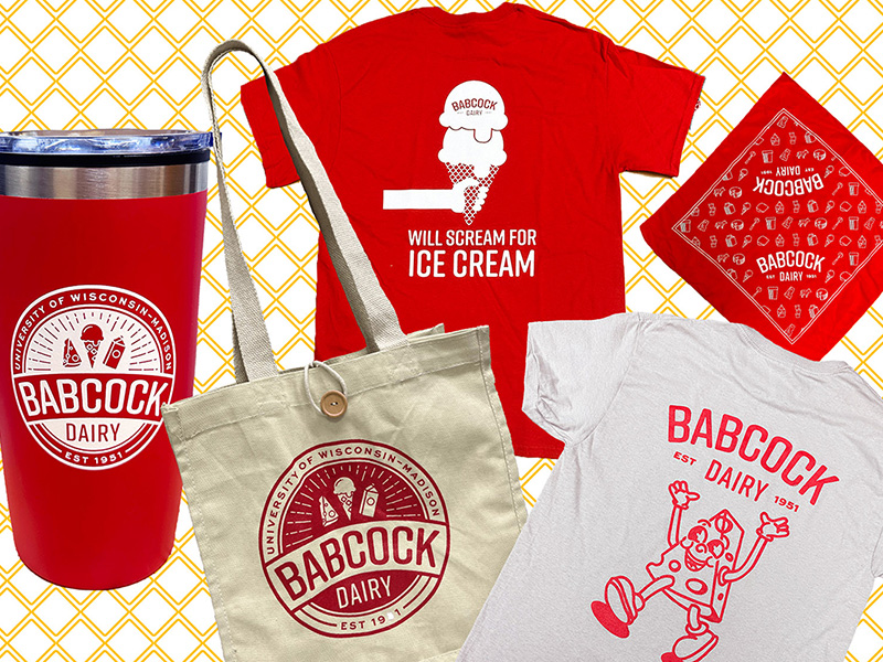 A collection of Babcock Dairy Store merchandise including a red travel tumbler, a beige tote bag, a red T-shirt with an ice cream graphic and the text ‘Will Scream for Ice Cream,’ a red bandana, and a light gray T-shirt with a cheese cartoon character, all featuring Babcock Dairy branding.