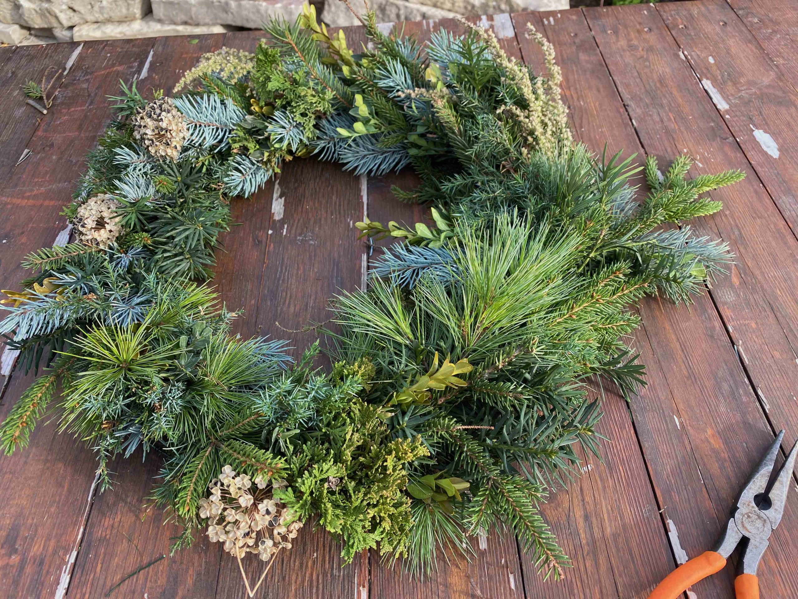 A completed wreath made of assorted evergreen branches and dried flowers arranged on a wooden table, with pruning shears placed nearby.
