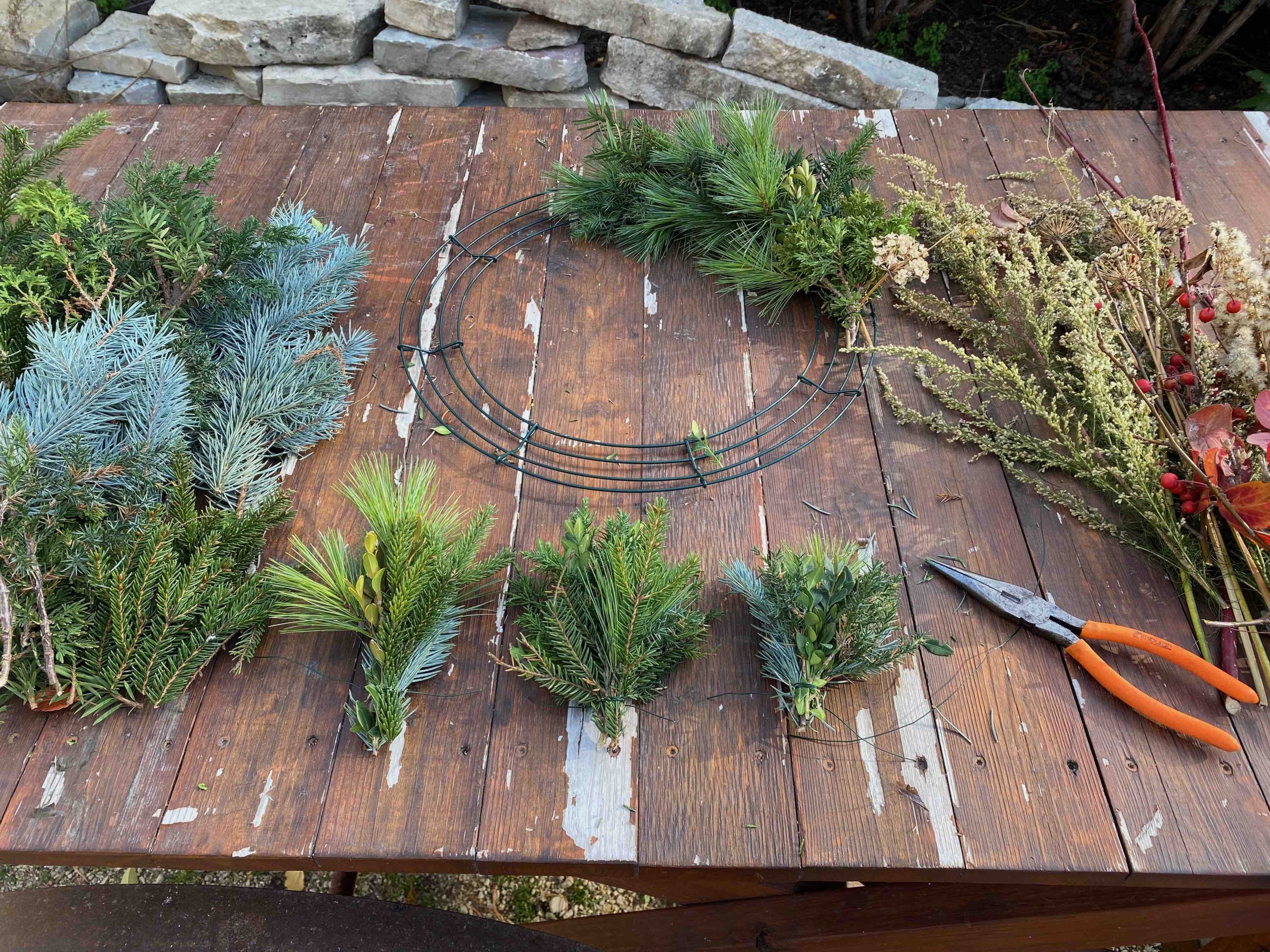 A wooden table displaying materials for making a wreath, including assorted evergreen branches, a metal wreath frame, floral wire, pruning shears, and dried plants with red berries.