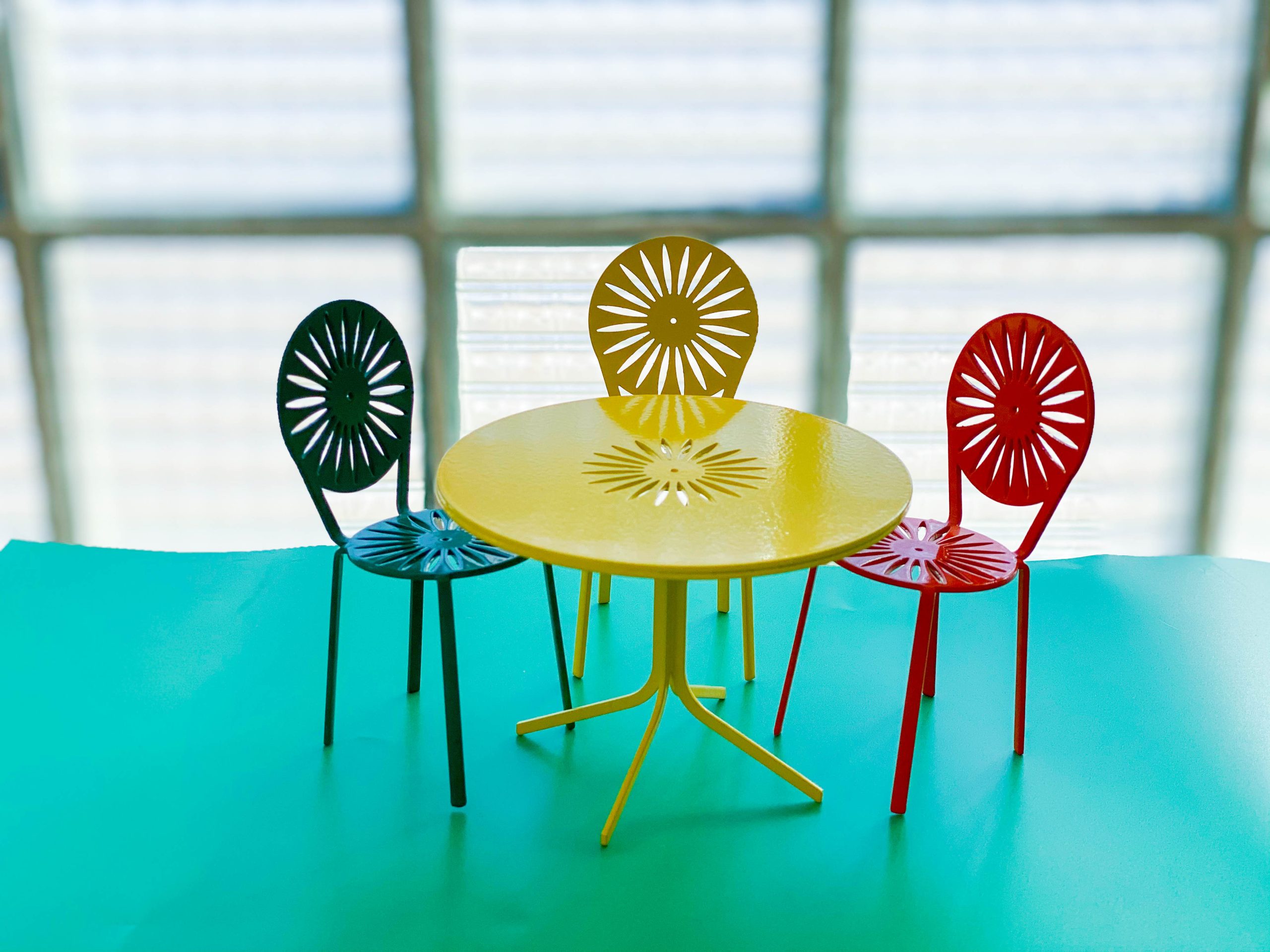 Three miniature terrace chairs in green, yellow, and red with sunburst-pattern backs surround a matching yellow round table on a turquoise surface, with a blurred glass block wall in the background.