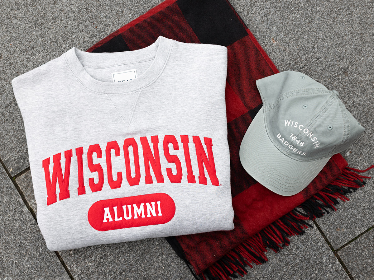 A folded gray sweatshirt with red text reading ‘Wisconsin Alumni’ placed on a red-and-black plaid blanket next to a light gray baseball cap with white text reading ‘Wisconsin Badgers, 1848’.