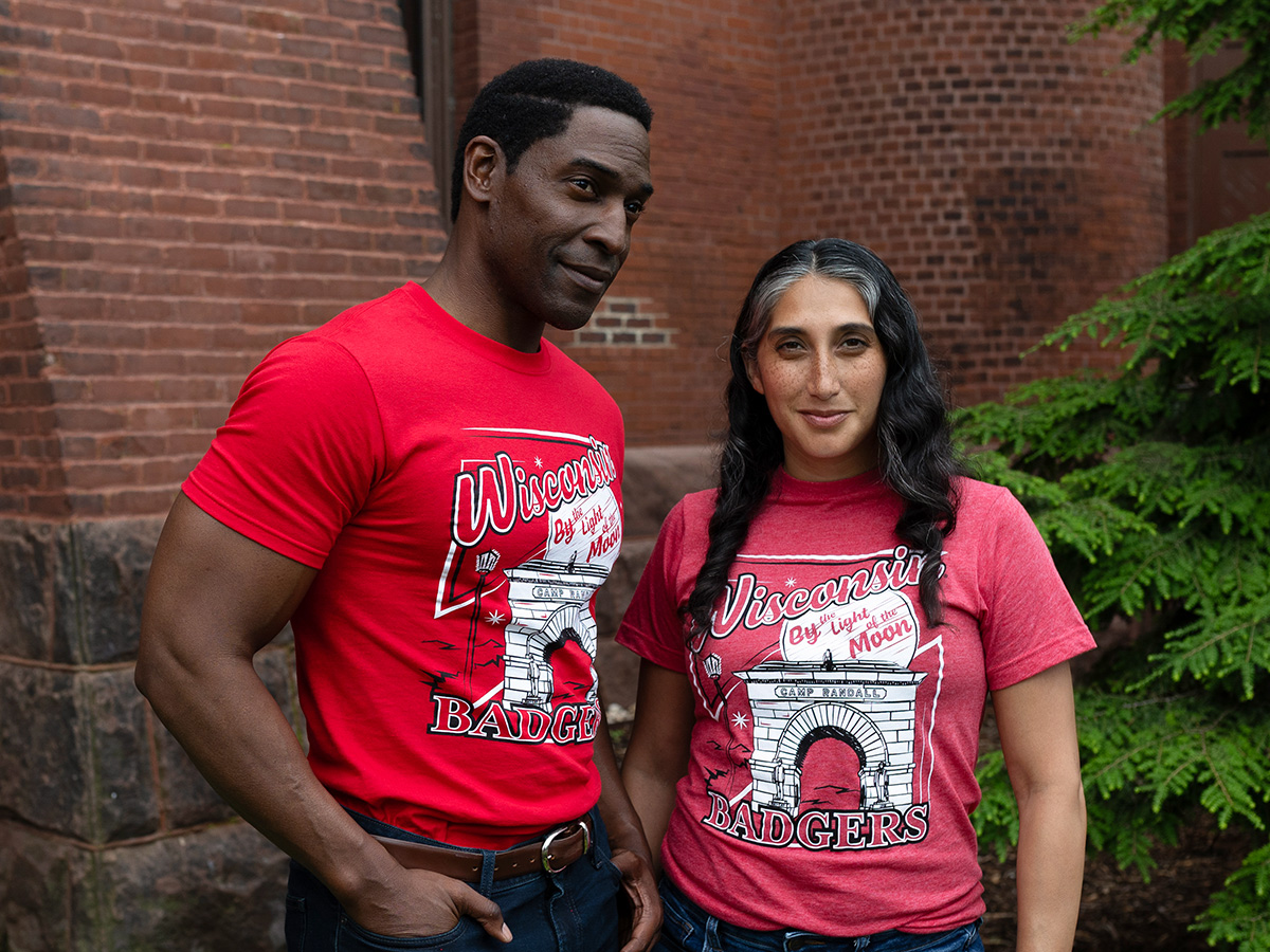 Two people standing outdoors in front of a brick wall and greenery, both wearing red Wisconsin Badgers T-shirts featuring an illustration of the Camp Randall Memorial Arch and the text ‘Wisconsin Badgers' and 'By the Light of the Moon'.
