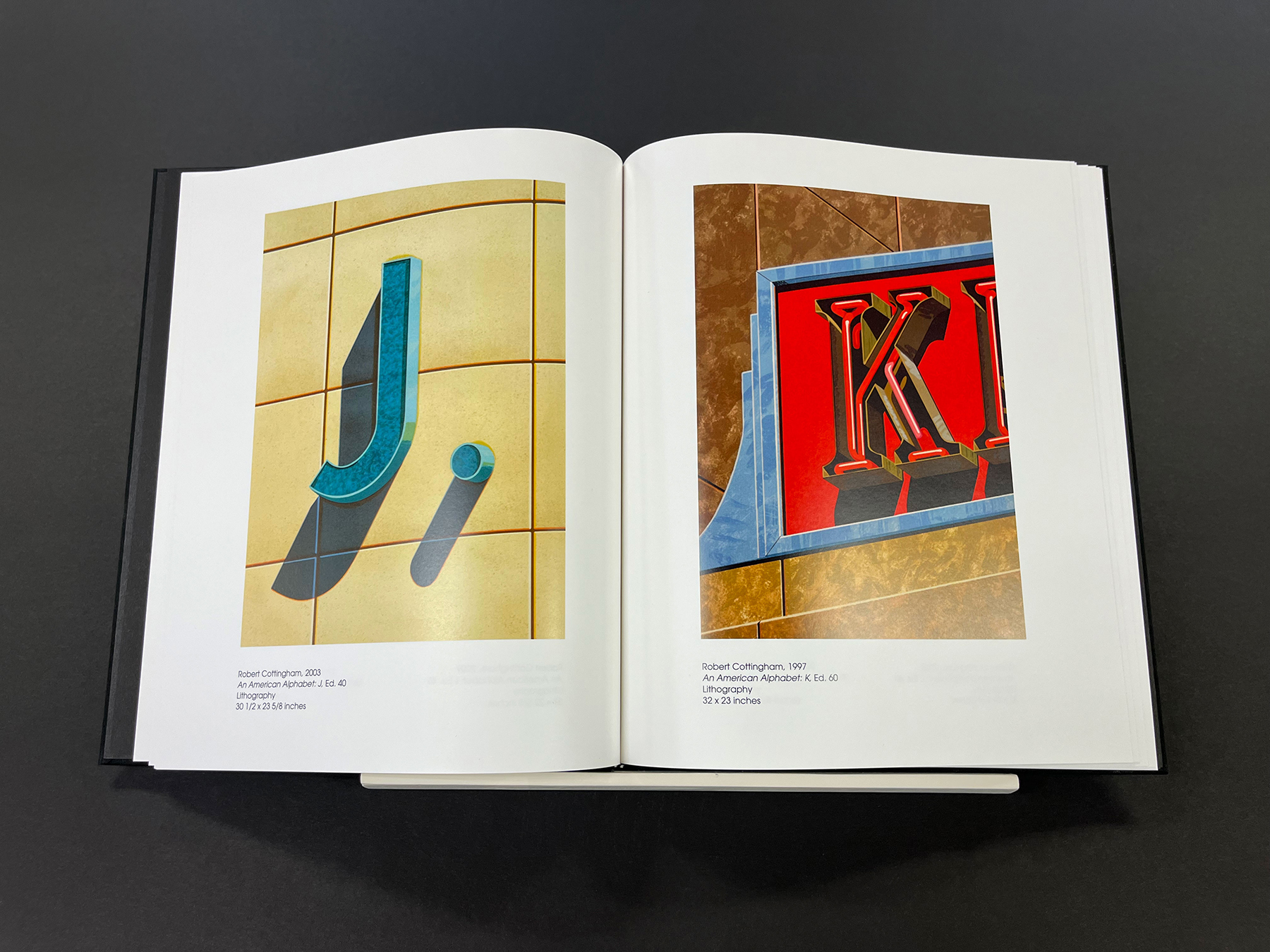 Open book showing two colorful artworks of letters: the left page features a blue letter ‘J’ with a period on a yellow tiled background, and the right page shows the letter ‘K’ on a bright red panel with blue and brown background elements.