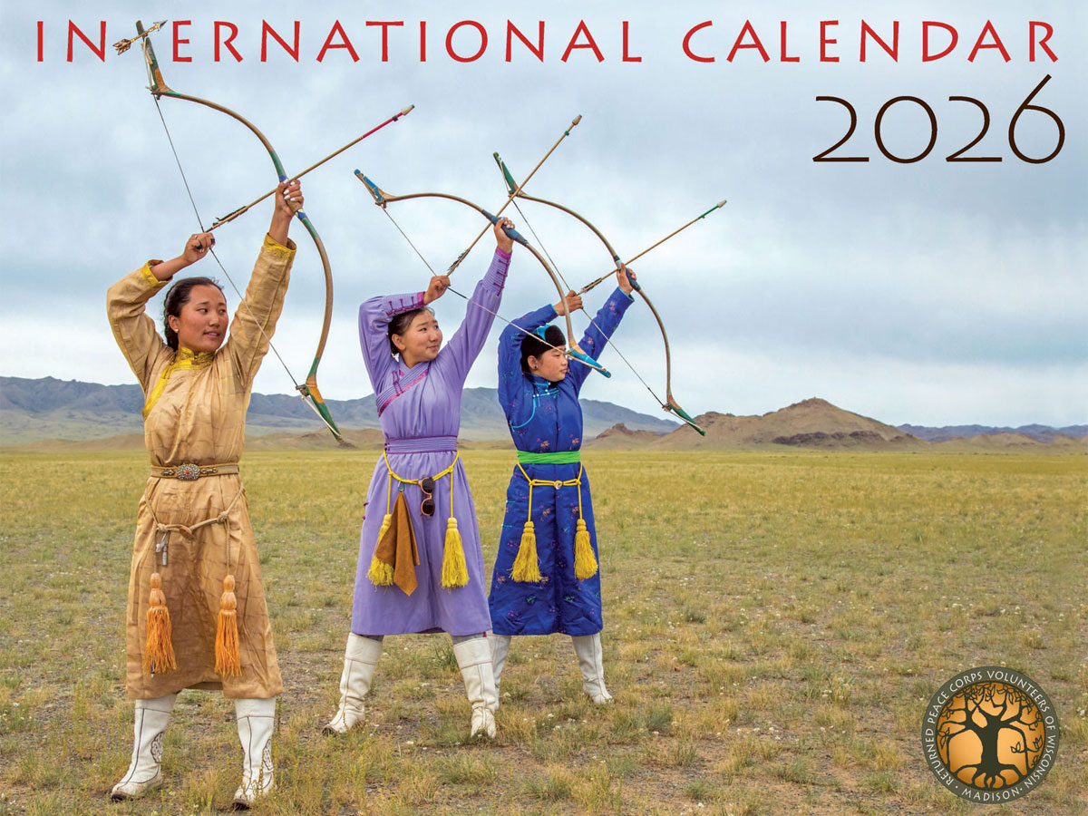 Cover of the 2026 International Calendar showing three people in colorful traditional outfits standing on a grassy plain and raising bows toward the sky, with mountains in the distance.