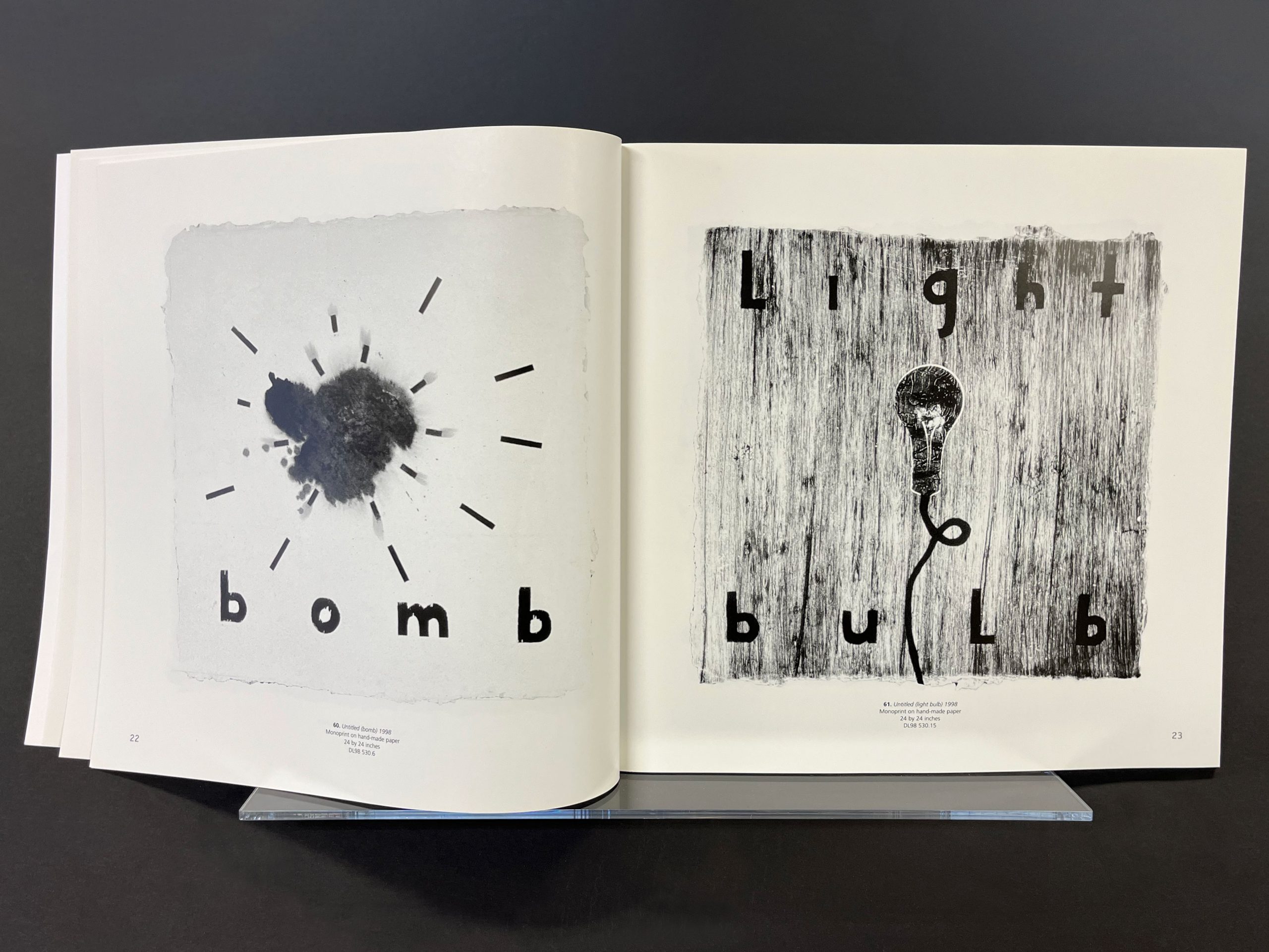 Open book displaying two monochromatic prints: the left page shows an abstract dark blot with radiating lines and the word ‘bomb,’ while the right page depicts a textured background with a light bulb illustration and the words ‘Light bulb.’