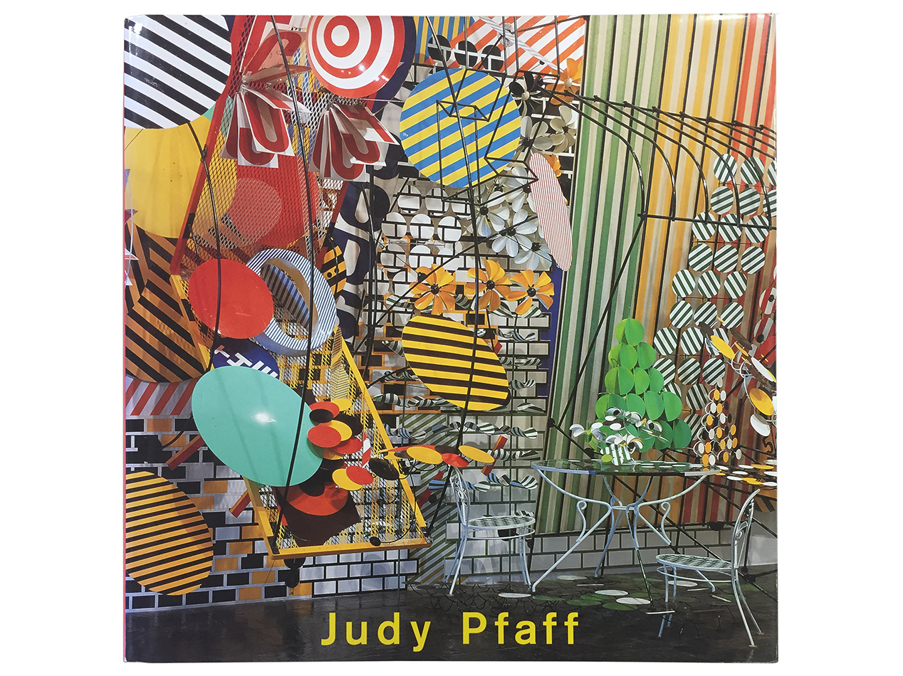 Cover of a book titled ‘Judy Pfaff’ featuring a vibrant abstract installation with colorful circular and striped shapes, wire structures, and metal chairs against a patterned background.