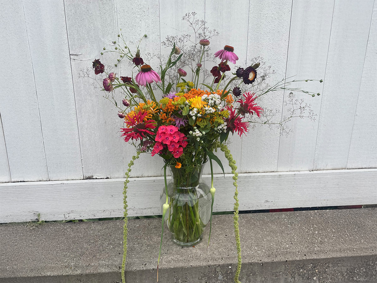 A clear glass vase filled with a tall, loose arrangement of wildflowers in pink, orange, yellow, white, and purple against a white wooden background.
