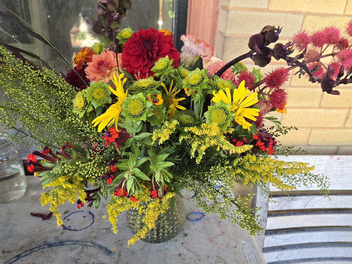 A textured glass vase holding a vibrant bouquet of mixed flowers and green foliage on a table near a brick wall.