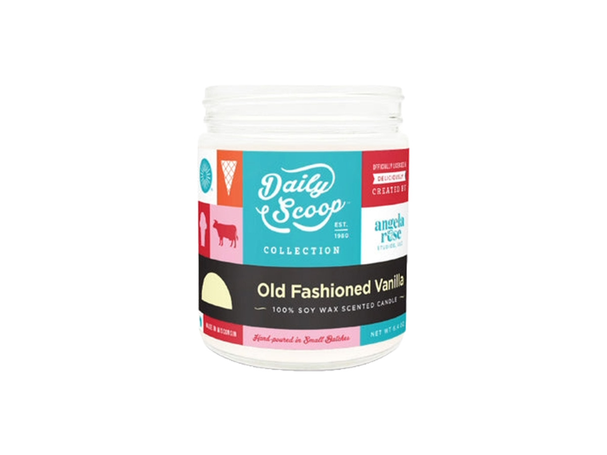 A white jar candle labeled 'Daily Scoop Collection Old Fashioned Vanilla' with colorful graphics including an ice cream cone, cow silhouette, and terrace chair, indicating it is a 100% soy wax scented candle hand-poured in small batches.