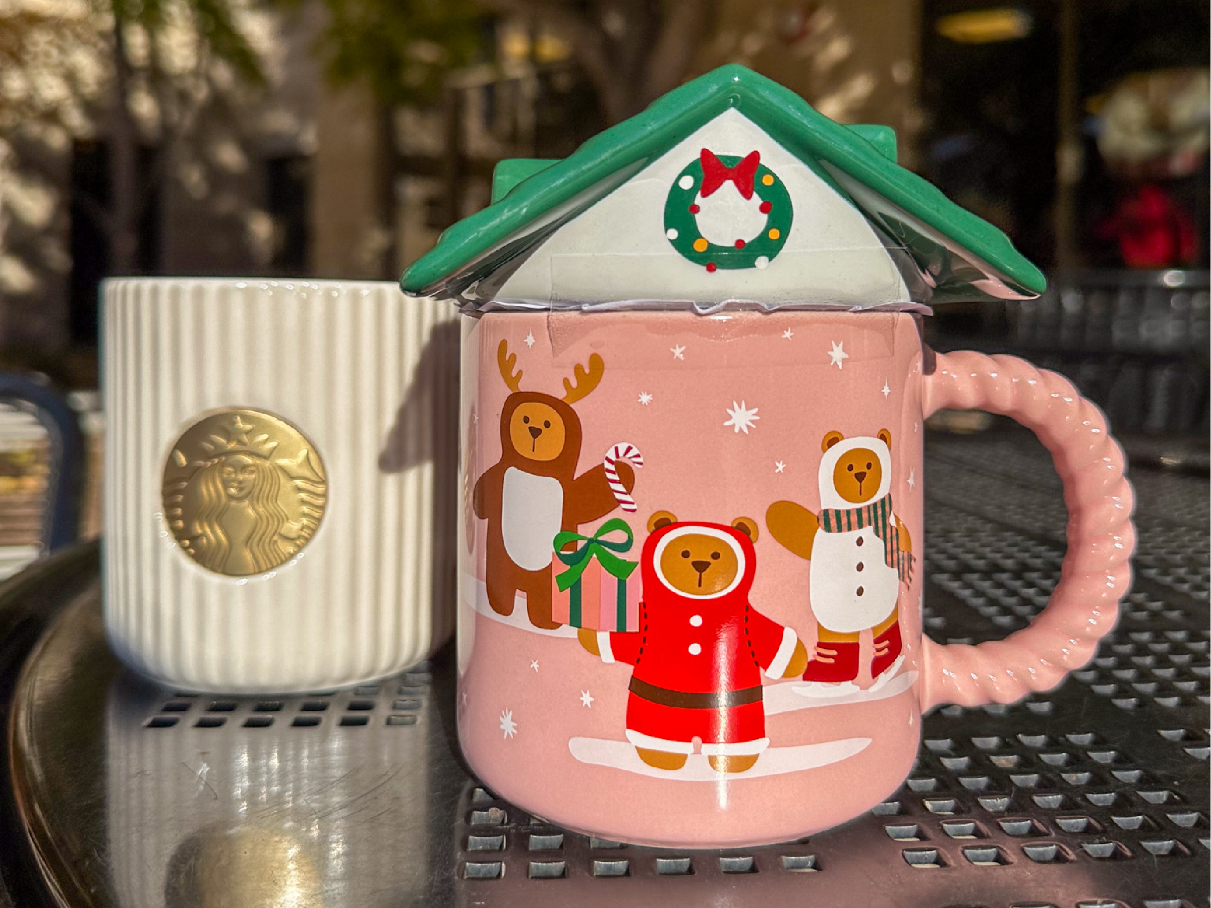 Two festive mugs sit on an outdoor metal table --one pink with holiday bear illustrations and a house-shaped lid, the other white with ridges and a gold Starbucks medallion -- capturing a cheerful winter theme.