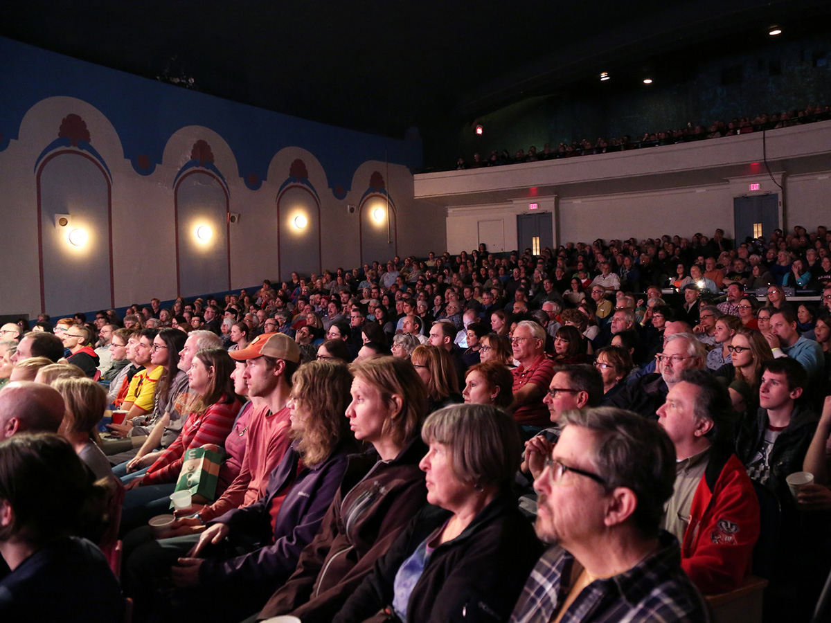 Large audience seated in a historic theater with ornate wall designs, watching a film under dim lighting.