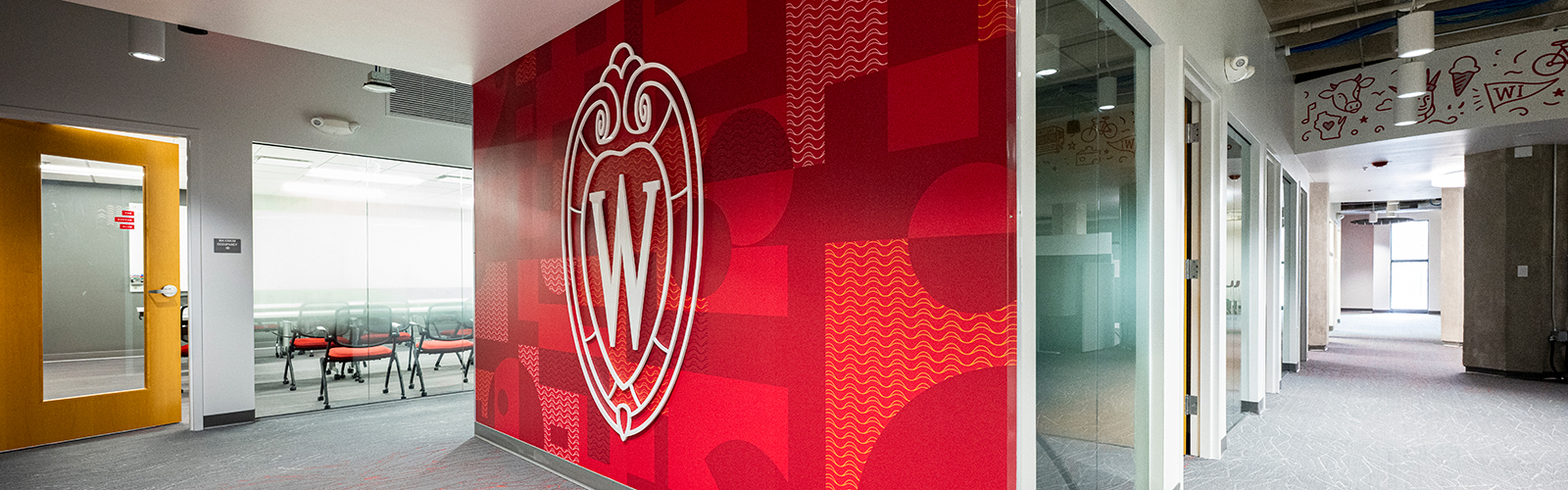 An office space showing a conference room with chairs, a large red entry wall with the UW–Madison crest logo, and a corridor
