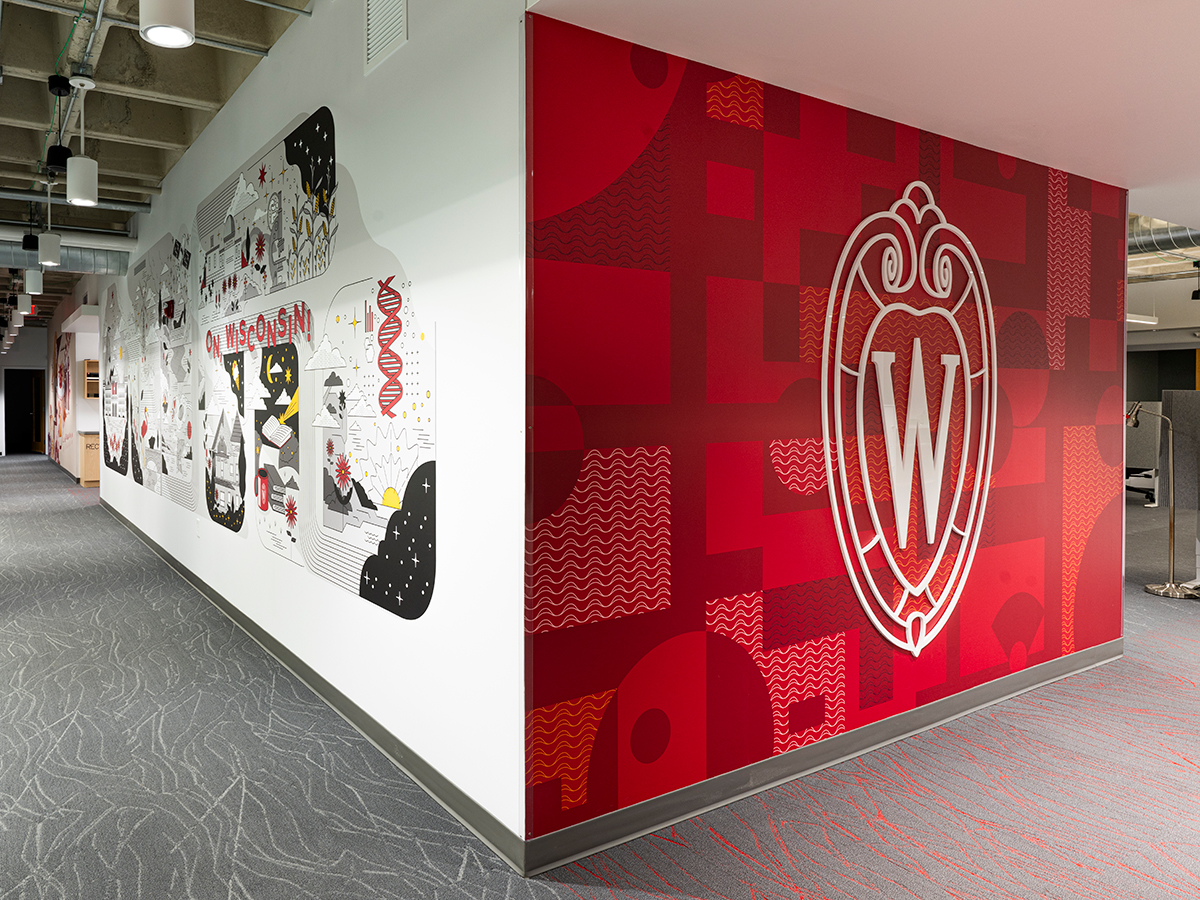 After photo of a red entry wall in a office setting with branded patterned wall paper with a UW–Madison dimensional crest logo on the wall.