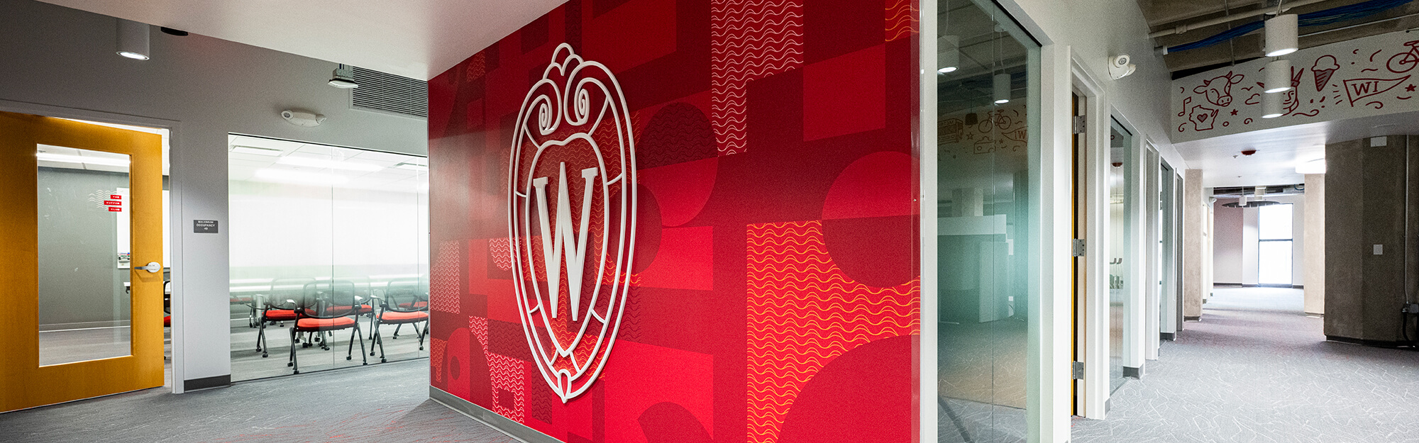 An office space showing a conference room with chairs, a large red entry wall with the UW–Madison crest logo, and a corridor.