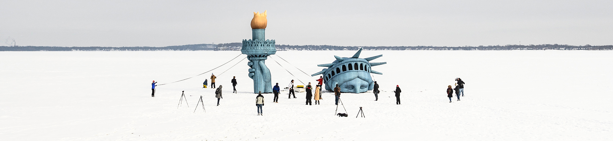 People stand on a snowy lake beside large inflatable pieces of the Statue of Liberty’s crown and torch.