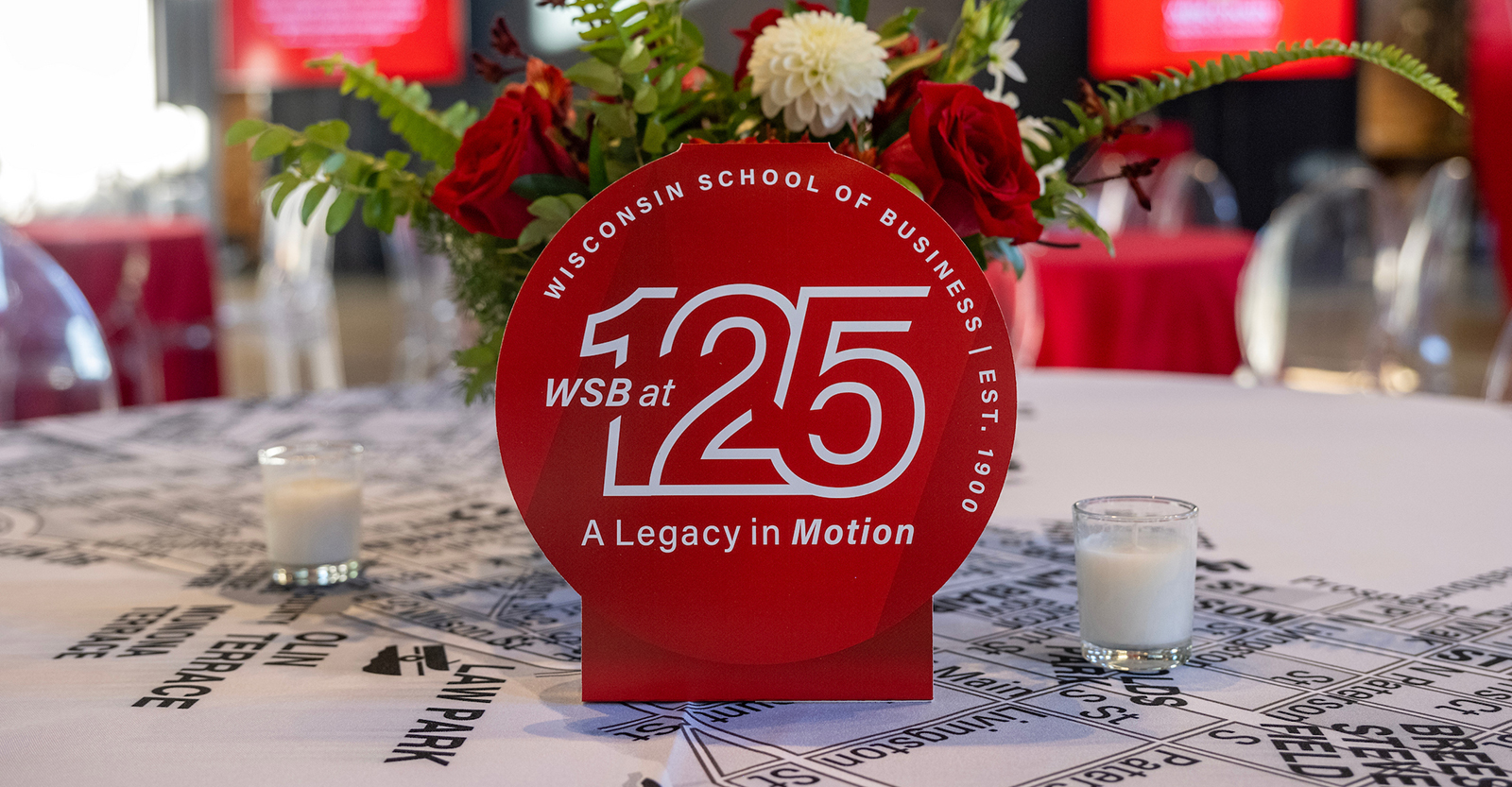 A Wisconsin School of Business 125th anniversary table tent on a table with flowers behind it.
