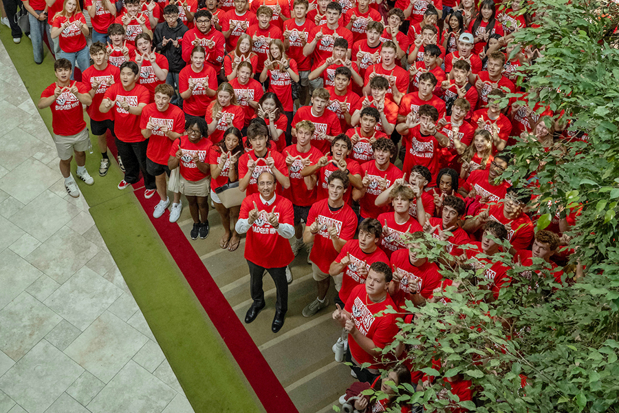 Large group of people wearing red shirts, making W sign with their hands, looking up and smiling at the camera.