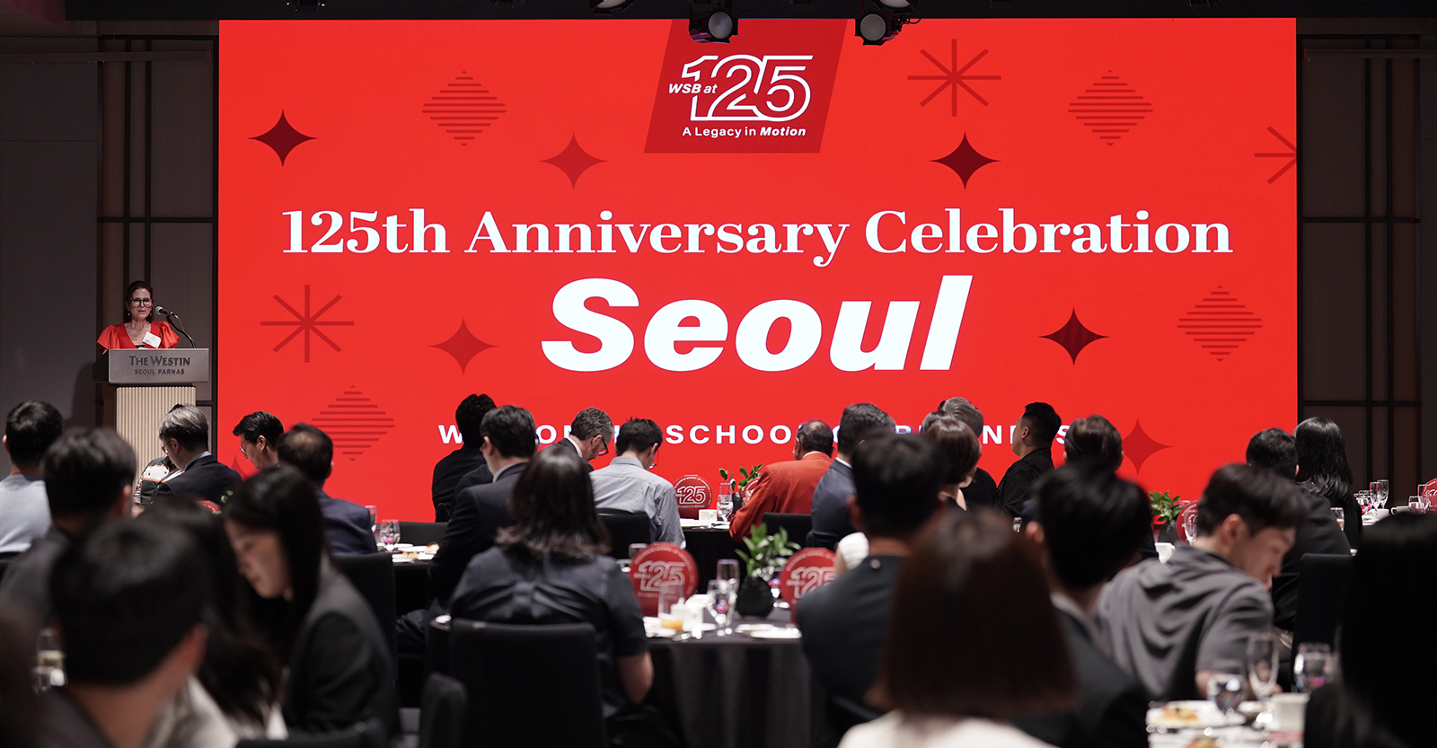 A speaker at a podium with people sitting at tables and a large screen with a slide that reads "125th Anniversary Celebration Seoul; Wisconsin School of Business"
