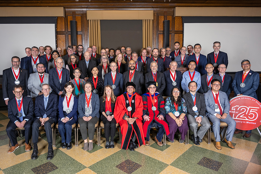 Large group of people in business attire wearing medals around their necks, smiling at the camera with a Wisconsin School of Business 125th anniversary sign on the right.