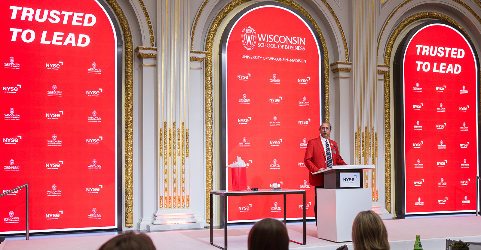 A person speaking at a podium with three arches in the background showing Wisconsin School of Business branding.